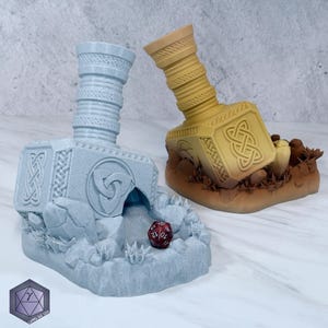 May include: Two decorative dice holders shaped like Thor's hammer, one light blue and one tan. The light blue holder has a red die inside. Both feature intricate Celtic knot designs and are set on a rocky base.