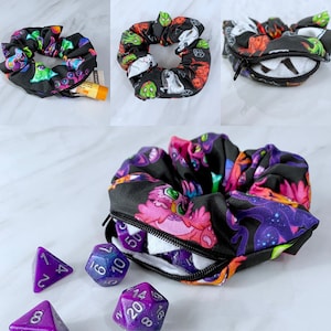 May include: A black scrunchie with a zippered pocket and a colorful print of cartoon monsters. The scrunchie is open and there are purple dice inside.