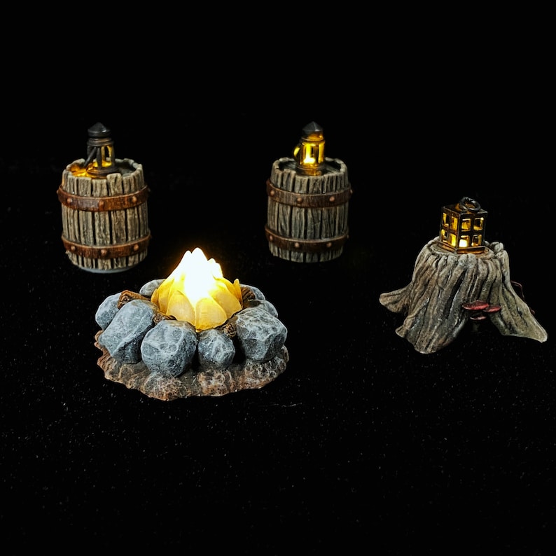 Puede incluir: Una escena de mesa miniatura con una fogata encendida rodeada de piedras, dos barriles de madera con linternas y un toc&oacute;n de &aacute;rbol con una linterna en la parte superior. Las linternas est&aacute;n encendidas y emiten un brillo c&aacute;lido.