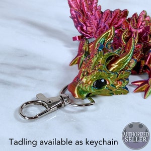 May include: A small, iridescent, green and pink dragon keychain with a silver metal clasp. The dragon has a detailed, textured design and a black eye. The keychain is attached to a silver metal ring.