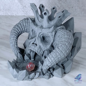 May include: Gray 3D printed dice tower shaped like a skull with horns and a crown. The skull is holding a red 20-sided die in its mouth.