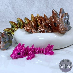 May include: Two 3D printed crystal dragon figurines. The larger dragon is brown, gold, and green. The smaller dragon is pink. Both dragons are lying on a white ceramic dish.