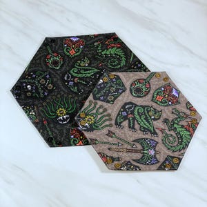 May include: Two hexagonal fabric coasters. One is dark gray with green, red, and purple fantasy-themed illustrations. The other is tan with similar illustrations. Both have gold-colored snaps around the edges.