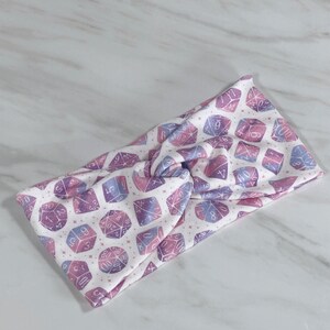 Puede incluir: Una diadema blanca con un patr&oacute;n de dados rosa y morado. Los dados tienen n&uacute;meros blancos.