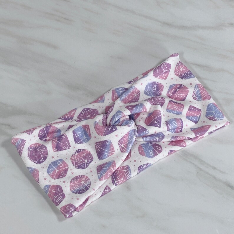Puede incluir: Una diadema blanca con un patr&oacute;n de dados rosa y morado. Los dados tienen n&uacute;meros blancos.
