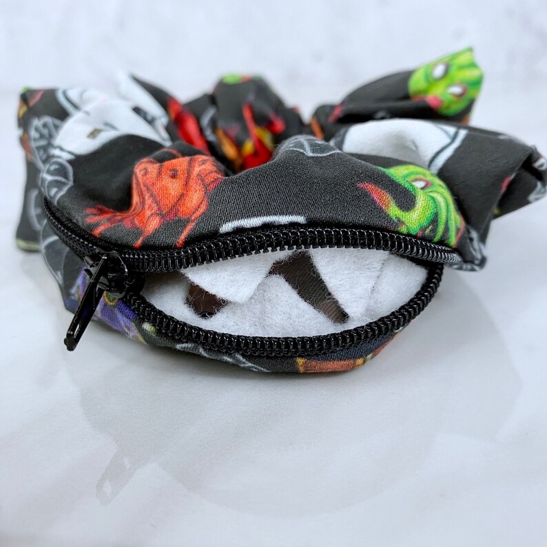May include: A black and white fabric scrunchie with a zipper closure. The scrunchie has a colorful print of various Halloween characters, including ghosts, monsters, and pumpkins.