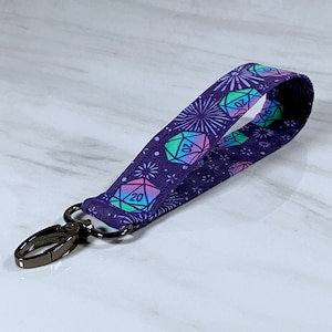 May include: A purple key chain with a repeating pattern of rainbow coloured 20-sided dice and fireworks. The key chain has a silver metal clip.