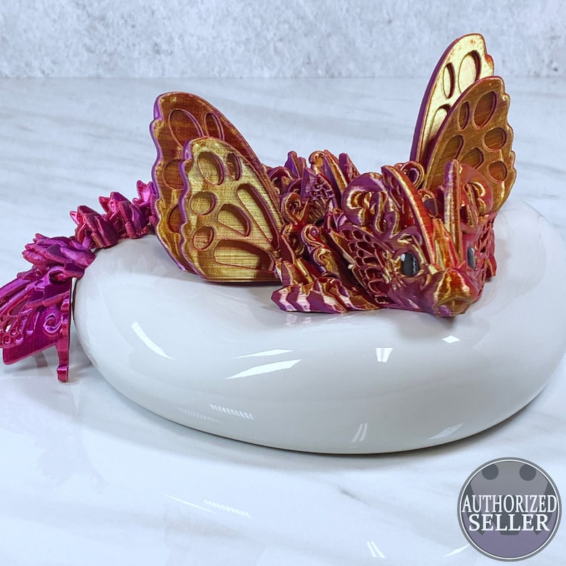 May include: A 3D printed dragon figurine with intricate details, featuring a red, gold, and pink color scheme. The dragon has large wings and a detailed face. The figurine is sitting on a white ceramic base.