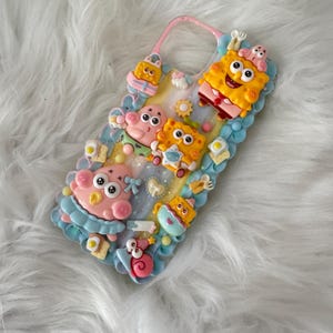 May include: A decorative phone case featuring a pastel blue whipped cream texture border and various 3D resin charms including Spongebob Squarepants, Patrick Star, a pink snail, small fried eggs on toast, and tiny cutlery pieces on a glittery base.