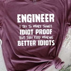 May include: Maroon t-shirt with the text "ENGINEER I TRY TO MAKE THINGS IDIOT PROOF BUT THEY KEEP MAKING BETTER IDOTS" in white. A white mug and flowers are next to the shirt.