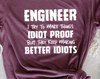 May include: Heather maroon t-shirt with the text "ENGINEER I TRY TO MAKE THINGS IDIOT PROOF BUT THEY KEEP MAKING BETTER IDIOTS" in white. A white mug and flowers are next to the shirt.