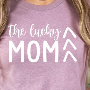 May include: Heather orchid-colored t-shirt with the text "the lucky MOM" in white. The shirt has a crew neck and short sleeves. The model is wearing blue denim shorts. The shirt is made of a soft, comfortable material.