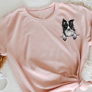 Könnte beinhalten: Ein T-Shirt in der Farbe Heidelbeer-Pfirsich mit einem schwarz-weißen Border Collie, der aus der Tasche schaut. Das Shirt liegt flach auf einer weißen Oberfläche mit einer weißen Decke und Blumen.