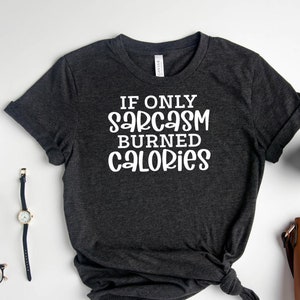 May include: A dark grey t-shirt with the text "If only sarcasm burned calories" in white lettering. The shirt is laid flat on a white surface with a coffee mug, a small brown purse, a watch, a pair of glasses, and a small black tray with gold earrings.