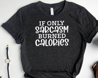 以下が含まれることがあります： 白い文字で「If only sarcasm burned calories」と書かれた濃いグレーのTシャツ。白い背景に、コーヒーカップ、小さな茶色のハンドバッグ、腕時計、眼鏡、そして金色のイヤリングが入った小さな黒いトレイが置かれている。