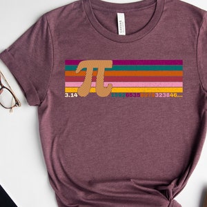 May include: Heather maroon t-shirt featuring a colorful Pi symbol design with the number 3.14 and a sequence of numbers. The design includes a series of horizontal stripes in various colors. The shirt is styled with accessories.