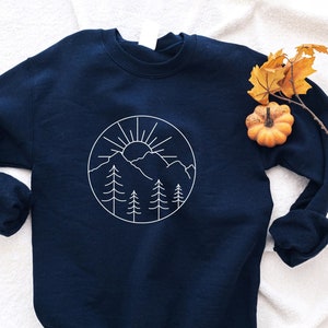 K&ouml;nnte beinhalten: Ein marineblauer Pullover mit einem wei&szlig;en Umriss einer Bergkette, Sonne und B&auml;umen in einem Kreis auf der Vorderseite. Der Pullover liegt auf einer wei&szlig;en, flauschigen Oberfl&auml;che.