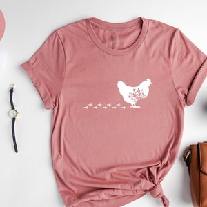 May include: Heather mauve t-shirt with a white graphic of a chicken with floral details. The chicken is walking with a trail of footprints behind it. The shirt is laid flat on a white surface.