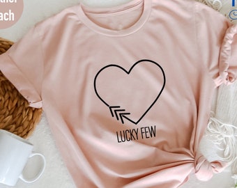 May include: A heather peach t-shirt with a black heart and arrows pointing to it. The text "Lucky Few" is below the heart.