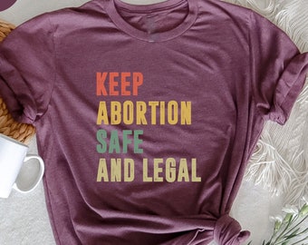 Könnte beinhalten: Ein T-Shirt in Heide-Maroon mit dem Text "KEEP ABORTION SAFE AND LEGAL" in einer Vintage-Schriftart mit bunten Akzenten.