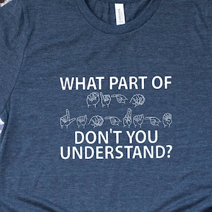 May include: A blue t-shirt with the text "WHAT PART OF DON'T YOU UNDERSTAND?" in white lettering. The text is accompanied by a row of hand signs in sign language. The shirt is paired with denim shorts, a brown leather handbag, and sparkly silver flats.
