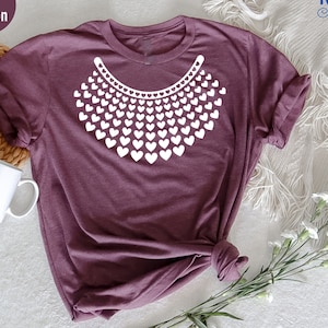 May include: A heather maroon t-shirt with a white graphic of a necklace made of hearts. The graphic is a tribute to Ruth Bader Ginsburg.