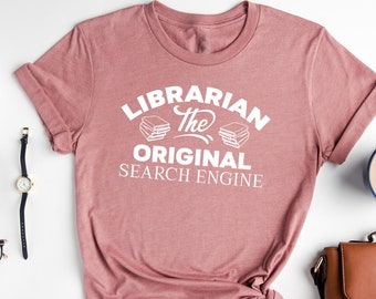 May include: Heather mauve t-shirt with the text "Librarian the original search engine" in white. The shirt is folded and tied in a knot. A brown leather purse, a cup of coffee, and a pair of glasses are also in the image.