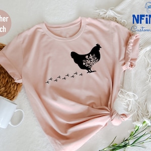 May include: A heather peach t-shirt with a black silhouette of a chicken with floral details and a trail of footprints. The text "NFINITI Custom Shirts" is in the top right corner of the image.