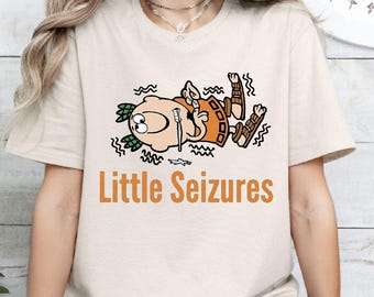 Puede incluir: Una camiseta de manga corta color arena con un personaje de dibujos animados tumbado en el suelo rodeado de líneas onduladas. El personaje lleva una túnica naranja y sandalias marrones. Debajo del gráfico, el texto Little Seizures está impreso en naranja.