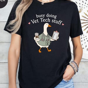 May include: A black t-shirt featuring a graphic of a white goose wearing a light green scrub top with a stethoscope, holding a coffee cup and a clipboard labeled Patient List. The text above the goose reads busy doing Vet Tech stuff.