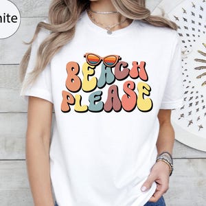 May include: White t-shirt with the text "BEACH PLEASE" in colorful, retro-style lettering. The design includes sunglasses above the word "BEACH". The shirt is a casual, everyday wear item.