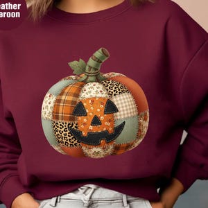 Op de afbeelding: Een heather maroon sweatshirt met lange mouwen en een centrale afbeelding van een pompoen gemaakt van verschillende stoffen lapjes. Het pompoenontwerp bevat ruitjes, bloemenprints en luipaardprints met een zwart jack-o'-lantern gezicht.