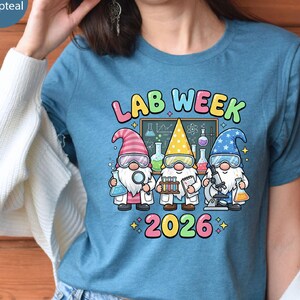 Pode incluir: Uma camiseta azul-petróleo com as palavras "LAB WEEK 2026" e três gnomos cientistas. Os gnomos usam jalecos de laboratório, óculos e chapéus coloridos. Um gnomo segura uma lupa, outro tubos de ensaio e o terceiro um microscópio.