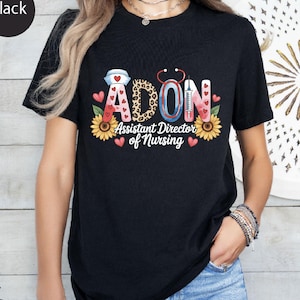 May include: A black t-shirt featuring a graphic design with the text ADON and Assistant Director of Nursing. The design includes a nurse cap, a stethoscope, a syringe, sunflowers, and small red hearts in a mix of pink, leopard, and blue colours.