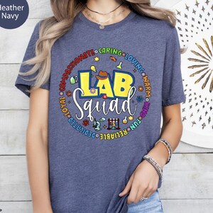 Pode incluir: T-shirt azul marinho mesclado com um gráfico colorido "Lab Squad". O design apresenta um balão de laboratório amarelo, um microscópio e outros ícones relacionados à ciência. As palavras "compassivo", "atencioso", "amoroso", "caloroso", "gentil", "divertido", "confiável", "dedicado" e "leal" circundam o texto principal.