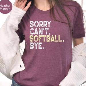 May include: A heather maroon short-sleeve t-shirt featuring white and yellow distressed text that reads SORRY. CAN'T. SOFTBALL. BYE. The shirt is worn with a white long-sleeve cardigan and blue denim jeans, showcasing a casual layered look.