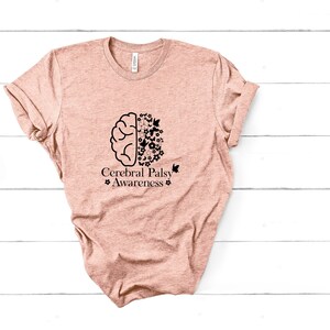 Może przedstawiać: Jasnor&oacute;żowy t-shirt z czarnym konturem m&oacute;zgu z kwiatami i motylami po jednej stronie i tekstem "Cerebral Palsy Awareness" po drugiej.