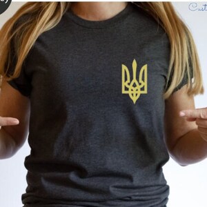 May include: A heather dark grey t-shirt with a gold embroidered Ukrainian trident symbol. The shirt is being modeled by a person. The text "NFINITI Custom Shirts" is visible in the top right corner of the image.