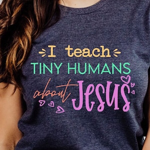 May include: A navy blue t-shirt with the text "I teach tiny humans about Jesus" in colorful lettering. The shirt has a crew neck and short sleeves. The shirt is made of a soft material.