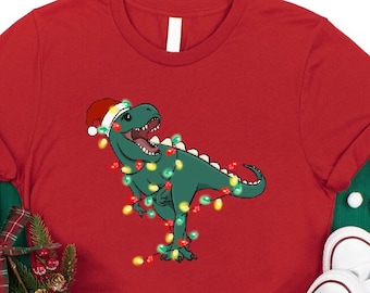 May include: Red t-shirt featuring a green dinosaur wearing a Santa hat and wrapped in Christmas lights. The shirt has the text "NFINITI Custom Shirts" in white. White sneakers and holiday decorations are in the background.