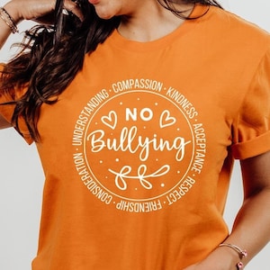 May include: Burnt orange t-shirt with a white circle graphic that says "No Bullying" with hearts and a floral design. The circle is surrounded by the words "Understanding, Compassion, Kindness, Acceptance, Respect, Friendship, Consideration".