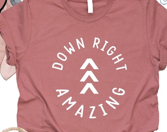May include: Heather mauve t-shirt with a white circle graphic that says "Down Right Amazing" with three white triangles inside the circle. The shirt is paired with denim shorts, a straw bag with a pink tassel, and white sandals.