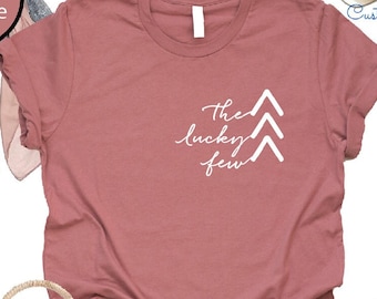 May include: A heather mauve t-shirt with the text "The lucky few" and three white arrows. The shirt is paired with denim shorts, a straw bag with a pink tassel, and white sandals.