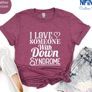 May include: Heather maroon t-shirt with the text "I love someone with Down Syndrome" in white. The shirt is shown on a white background with a pair of white shoes and denim shorts.