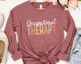 May include: A long-sleeved t-shirt in a mauve color with the text "Occupational Therapy" in white letters with a gradient background of orange, yellow, and brown.