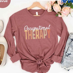 May include: A long-sleeved t-shirt in a mauve color with the text "Occupational Therapy" in white letters with a gradient background of orange, yellow, and brown.