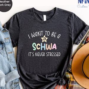 May include: A dark heather grey t-shirt with the text "I want to be a SCHWA it's never stressed" in colorful letters. The text is centered on the shirt.