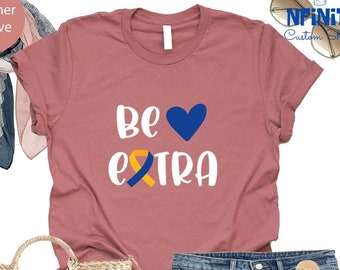Pode incluir: Camiseta heather malva com o texto "Be extra" em branco com um coração azul e uma fita amarela e azul. A camiseta é combinada com shorts jeans, uma bolsa de palha com uma borla rosa e sandálias brancas.