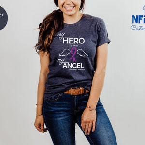 May include: Heather navy t-shirt with the text "my HERO is now my ANGEL" in white script, a purple ribbon, and angel wings. The shirt is from NFINITI Custom Shirts.