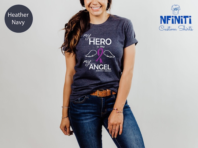 May include: Heather navy t-shirt with the text "my HERO is now my ANGEL" in white script, a purple ribbon, and angel wings. The shirt is from NFINITI Custom Shirts.